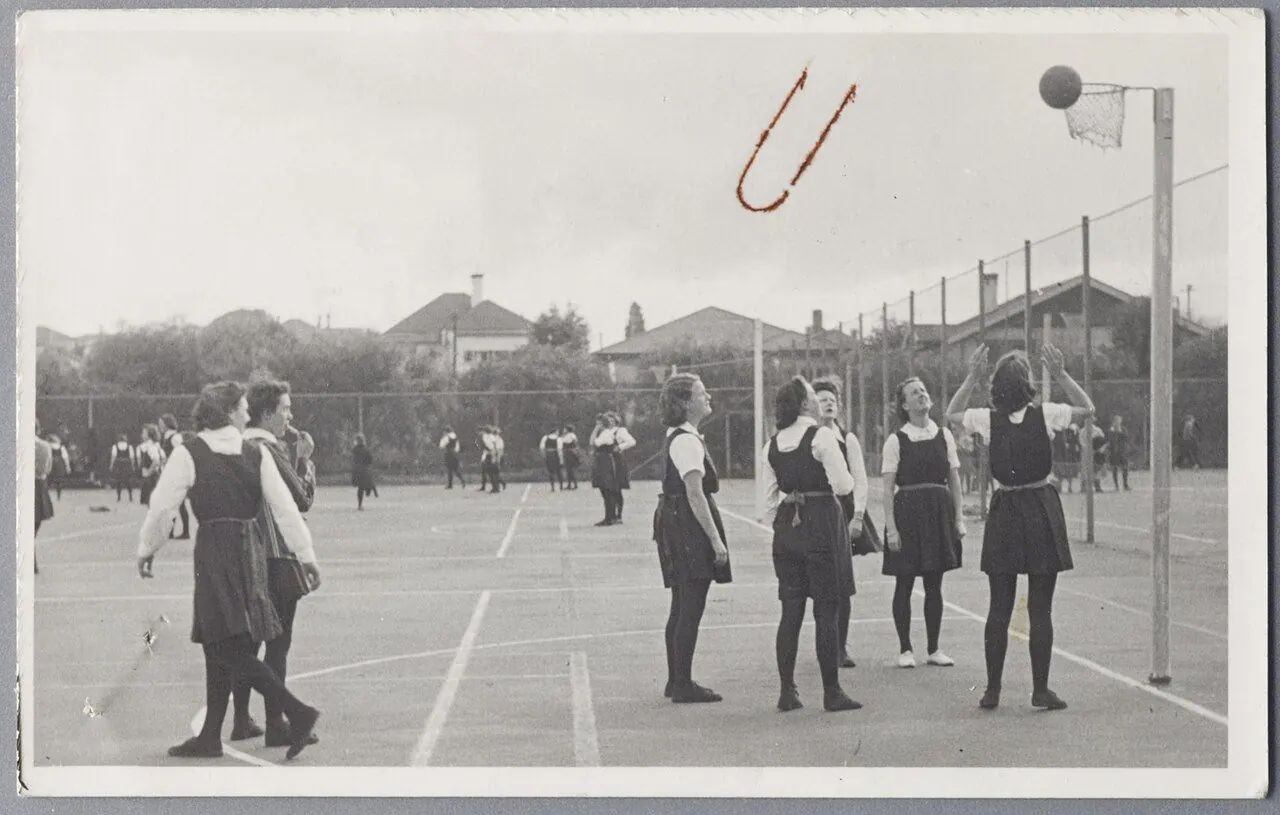 Scene from a netball encounter, Wellington. View looking at goalmouth action during a netball (or outdoor basketball) match - five players are looking transfixed at the ball as it makes contact with t